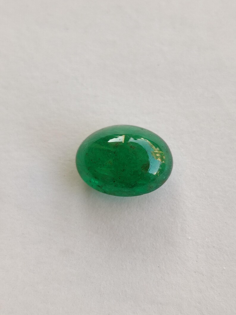 May include: A vibrant, oval-shaped, polished green gemstone. The gem has a smooth, glossy surface, reflecting light. The color is a rich, deep green, with subtle variations in tone. The gemstone is set against a plain, light background.