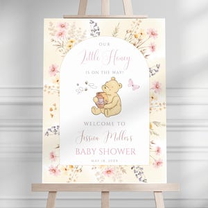 Puede incluir: Un letrero de bienvenida para baby shower con una ilustración de Winnie the Pooh. El letrero tiene un fondo floral y texto rosa que dice "Our Little Honey is on the Way!" y "Welcome to Jessica Millers Baby Shower". La fecha es el 18 de mayo de 20XX.