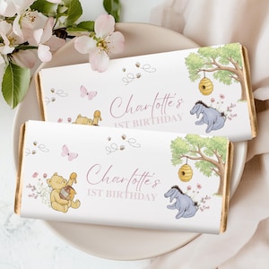 May include: Two chocolate bars with a Winnie the Pooh theme. The bars feature illustrations of Pooh, Eeyore, butterflies, and bees. The text "Charlotte's 1st Birthday" is written in a cursive font. The bars are on a white plate with pink flowers.