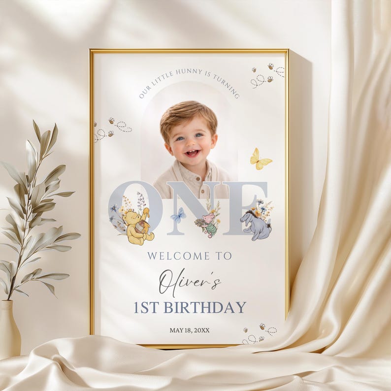 May include: A framed birthday sign with a photo of a smiling toddler. The sign reads "Our Little Hunny is Turning One" and "Welcome to Oliver's 1st Birthday" with the date May 18, 20XX. Winnie the Pooh characters and butterflies decorate the sign.