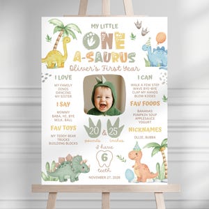 Könnte beinhalten: Ein dekoratives Poster auf einer Staffelei mit Dinosaurier-Motiv. Das Poster zeigt den Text "My Little One A-Saurus" und ein Foto eines Babys. Es enthält Informationen über das erste Lebensjahr des Babys, Lieblingsspielzeug und -nahrungsmittel. Das Baby wiegt 9.07 kg und ist 63.5 cm groß.