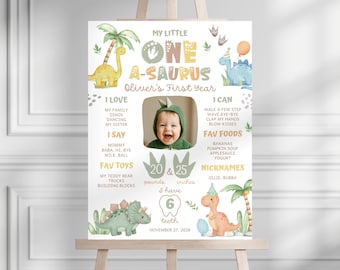 May include: A decorative poster on an easel with a dinosaur theme. The poster features the text "My Little One A-Saurus" and a photo of a baby. It includes information about the baby's first year, favorite toys, and foods. The baby is 20 pounds and 25 inches.