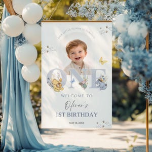 May include: A personalized birthday banner featuring a smiling child's photo, the word "ONE", and Winnie the Pooh characters. The banner reads "Our Little Hunny is Turning" and "Welcome to Oliver's 1st Birthday". Decorative balloons and floral accents surround the banner.