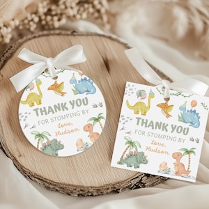 Può includere: Due etichette regalo a tema dinosauro con nastro bianco. Una è rotonda, l'altra quadrata. Entrambe presentano illustrazioni di dinosauri, palme e il testo "THANK YOU FOR STOMPING BY Love, Hudson."