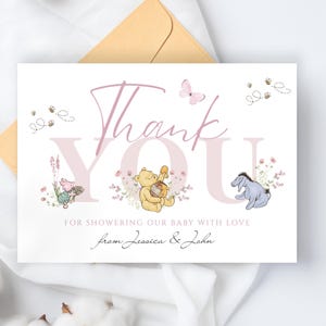 May include: A baby shower thank you card with a soft pink and beige color scheme. The card features illustrations of Winnie the Pooh characters, a butterfly, and bees. The text reads "Thank You" and "For showering our baby with love from Jessica & John."