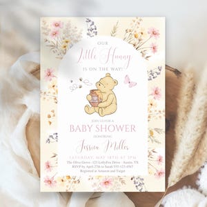 Puede incluir: Invitación a un baby shower con una ilustración de Winnie the Pooh. La invitación tiene un borde floral y el texto "Our Little Hunny is on the Way!" con detalles del evento, incluido el nombre de la homenajeada, la fecha, la hora y el lugar.
