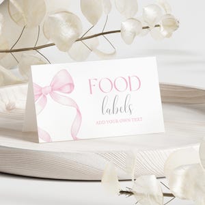 Könnte beinhalten: Eine weiße Essensetikettenkarte mit einer rosa Schleifenillustration und den Worten "FOOD labels" in rosa und schwarzer Schrift. Der Text "ADD YOUR OWN TEXT" steht unter dem Wort "labels". Die Karte liegt auf einem weißen Holztablett mit dekorativen weißen Blättern.
