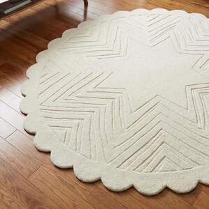 May include: Off-white, round rug with a large star design in the center. The star is made of zig-zag lines. The rug has a scalloped edge. The rug is on a hardwood floor.