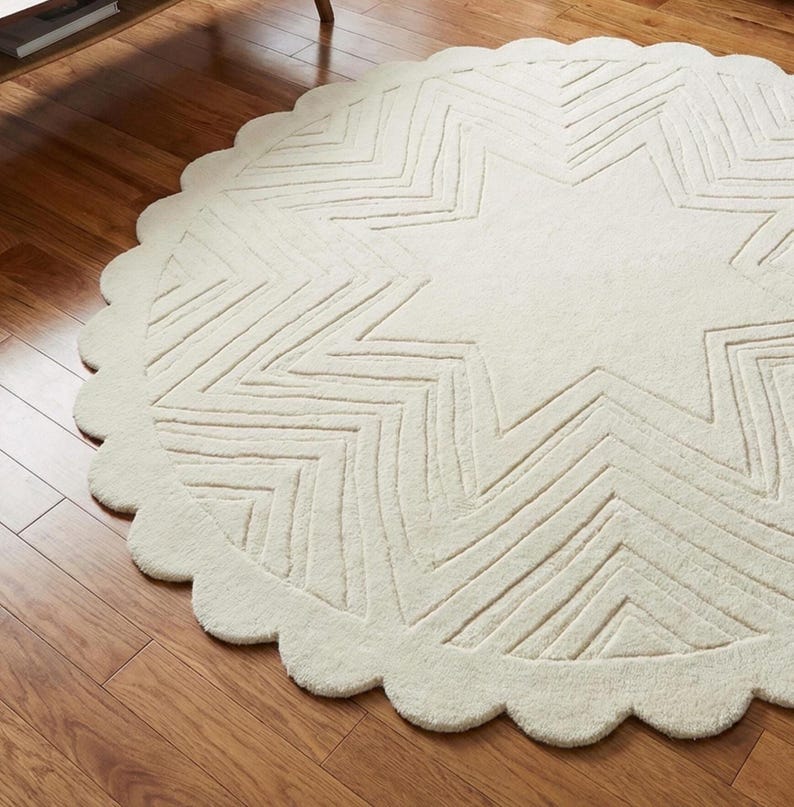 May include: Off-white, round rug with a large star design in the center. The star is made of zig-zag lines. The rug has a scalloped edge. The rug is on a hardwood floor.
