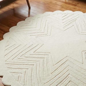 May include: Cream-colored, round rug with a star design. The rug features a scalloped edge and a large star pattern in the center, with zig-zag lines creating the star's points. The rug is made of a soft, textured material.