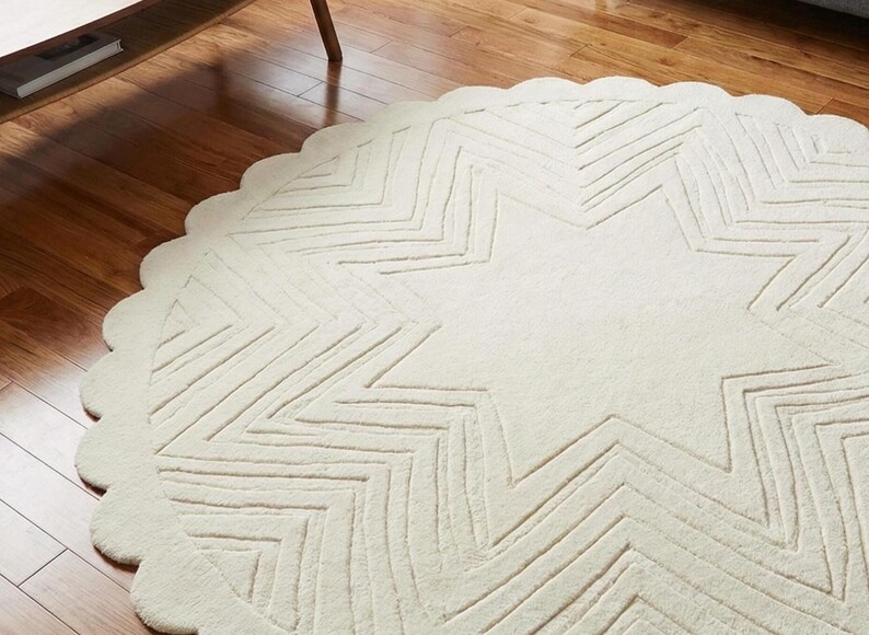 May include: Cream-colored, round rug with a star design. The rug features a scalloped edge and a large star pattern in the center, with zig-zag lines creating the star's points. The rug is made of a soft, textured material.