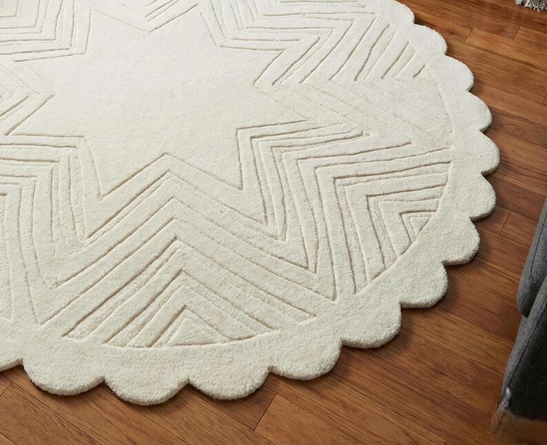 May include: Cream-colored, round rug with a star design. The rug features a scalloped edge and a textured surface. The star pattern is created with linear indentations, adding depth and visual interest to the floor covering.