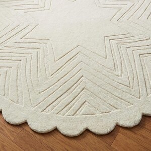 May include: Cream-colored, round rug with a starburst design. The rug features a scalloped edge and a large star pattern in the center, with zig-zag lines. The rug is placed on a hardwood floor.