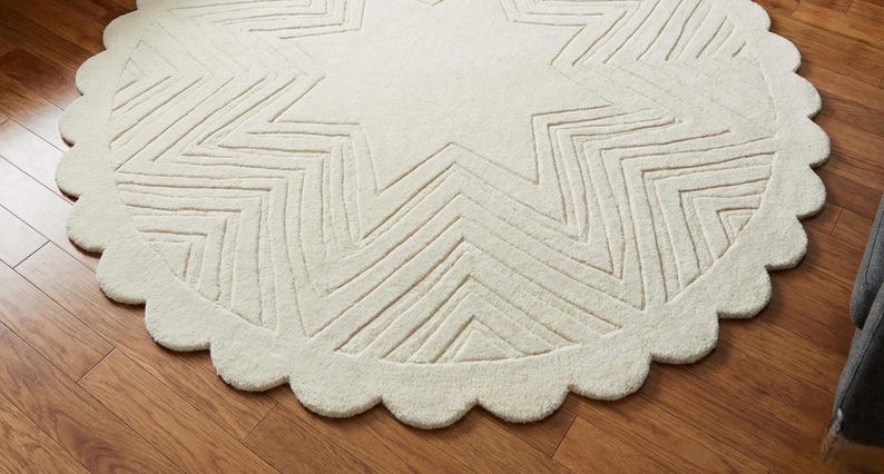May include: Cream-colored, round rug with a starburst design. The rug features a scalloped edge and a large star pattern in the center, with zig-zag lines. The rug is placed on a hardwood floor.