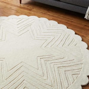 May include: Cream-colored, round rug with a large star design in the center. The star is made of zig-zag lines, and the rug has a scalloped edge. The rug is on a hardwood floor.