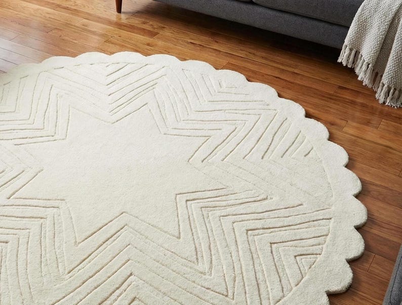 May include: Cream-colored, round rug with a large star design in the center. The star is made of zig-zag lines, and the rug has a scalloped edge. The rug is on a hardwood floor.