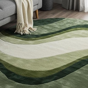 May include: A round area rug with a wave-like pattern in shades of green and white. The rug is made of a soft, plush material and is placed on a hardwood floor. The rug is near a gray sofa and a cream-colored throw blanket.