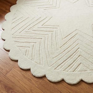 May include: Off-white, round rug with a star design. The rug has a scalloped edge and a textured surface. The star pattern is created with linear, zig-zag lines. The rug is made of a soft, plush material.