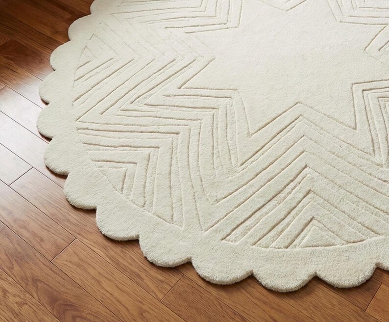 May include: Off-white, round rug with a star design. The rug has a scalloped edge and a textured surface. The star pattern is created with linear, zig-zag lines. The rug is made of a soft, plush material.
