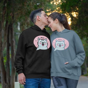 May include: Black and grey hoodies with a front pocket and a pink oval graphic of a cartoon cat with the word "Meow". The hoodies are part of a matching set. The image shows the hoodies being worn.