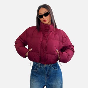 May include: A cropped burgundy puffer jacket with a high collar, long sleeves, and a front zipper closure. The jacket features horizontal quilted stitching throughout the body and sleeves, paired with blue denim jeans and black cat-eye sunglasses.