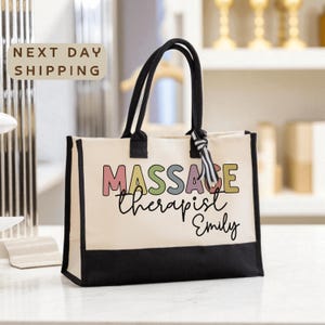 May include: Cream-colored tote bag with black accents and handles. The bag features the text "MASSAGE Therapist Emily" in colorful letters. A striped tag is tied to one of the handles. The bag is on a white surface.
