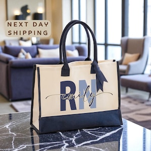 May include: Cream-colored tote bag with navy blue accents and handles. The bag features the initials "RN" in navy blue and the name "emily" in a script font. A navy blue ribbon is tied to the handle. The bag is sitting on a black marble surface.