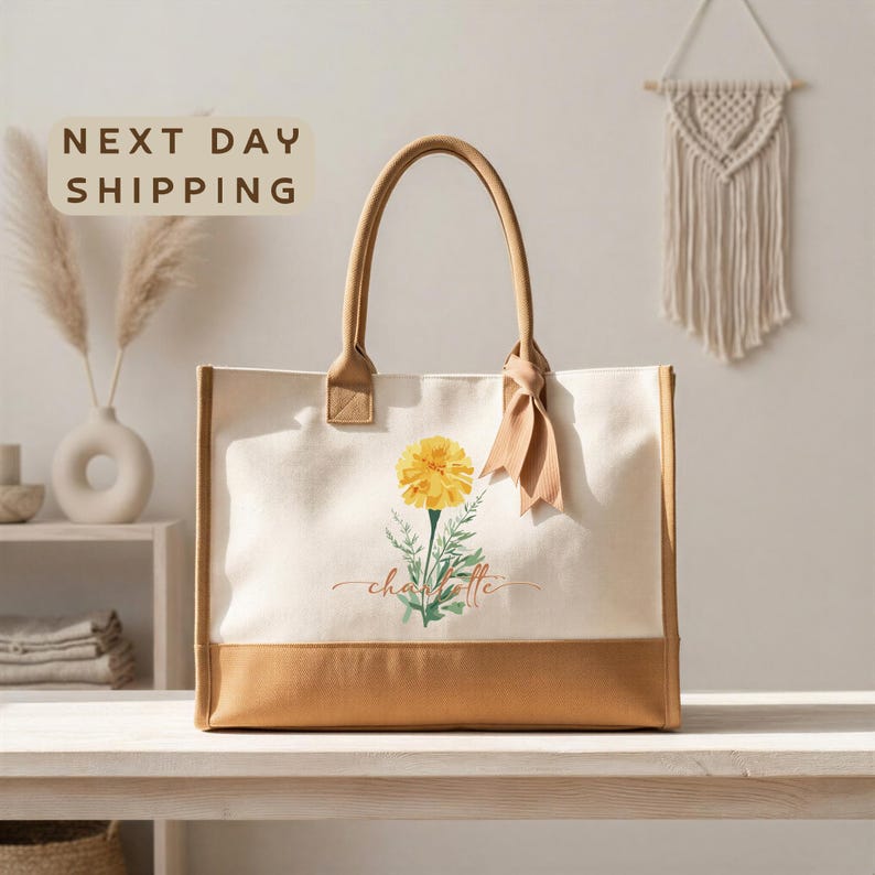 a beige tote bag with a yellow flower design, placed on a wooden surface. In the background, there is a wall with a macrame wall hanging and a shelf with various decorative items.