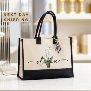 A beige tote bag with a black trim and a white lily of the valley design, featuring the name "Olivia" written in cursive. The bag is placed on a white surface, and there is a sign that says "Next day shipping" in the background.