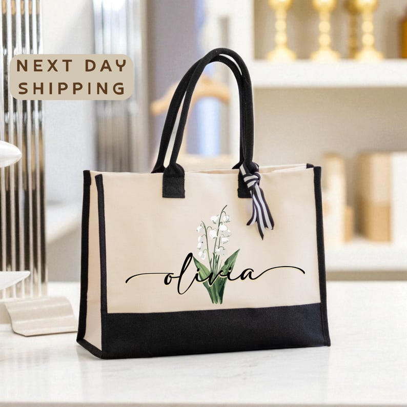 A beige tote bag with a black trim and a white lily of the valley design, featuring the name "Olivia" written in cursive. The bag is placed on a white surface, and there is a sign that says "Next day shipping" in the background.