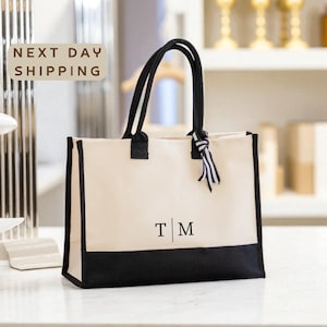 May include: Cream-colored tote bag with black accents and handles. The bottom of the bag is black, and the handles are black. The bag has the initials "T | M" printed on the front. A tag with black and white stripes is tied to the handle.