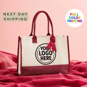 A white tote bag with a maroon trim, featuring a black circular logo that says "YOUR LOGO HERE". The bag is placed on a red fabric surface, and there is text in the background that reads "NEXT DAY SHIPPING FULL COLOR PRINTING".
