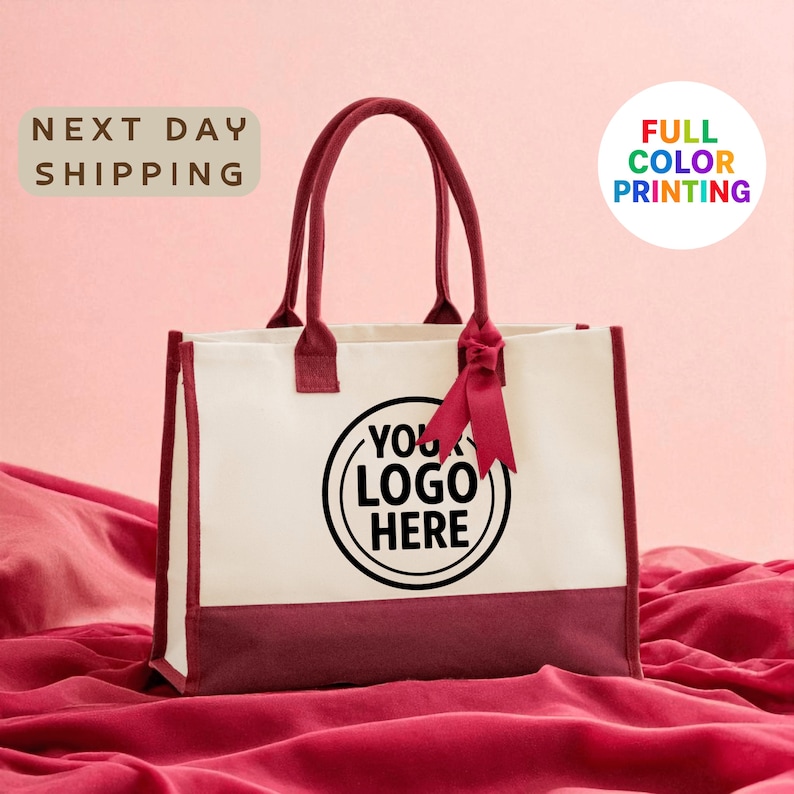 A white tote bag with a maroon trim, featuring a black circular logo that says "YOUR LOGO HERE". The bag is placed on a red fabric surface, and there is text in the background that reads "NEXT DAY SHIPPING FULL COLOR PRINTING".