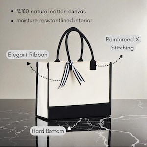 a white tote bag with black trim, placed on a marble surface. The bag has a black handle and a black and white ribbon tied around it. The bag is described as having a "Hard Bottom" and "Reinforced X Stitching", indicating its durability and sturdiness. The bag is also described as having "%100 natural cotton canvas" and "moisture resistant interior", suggesting that it is made of high-quality materials and can withstand wear and tear.