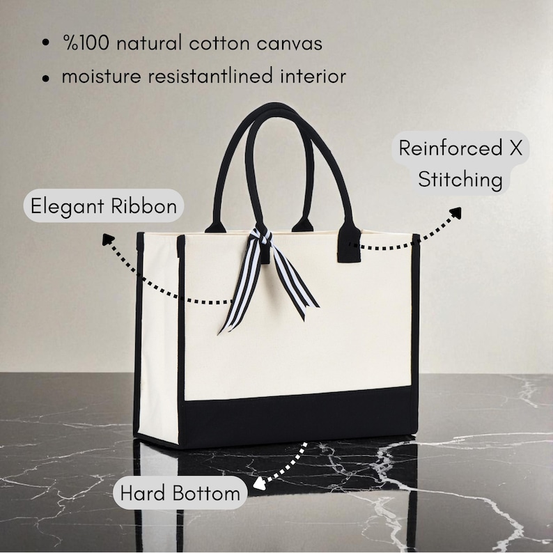 a white tote bag with black trim, placed on a marble surface. The bag has a black handle and a black and white ribbon tied around it. The bag is described as having a "Hard Bottom" and "Reinforced X Stitching", indicating its durability and sturdiness. The bag is also described as having "%100 natural cotton canvas" and "moisture resistant interior", suggesting that it is made of high-quality materials and can withstand wear and tear.