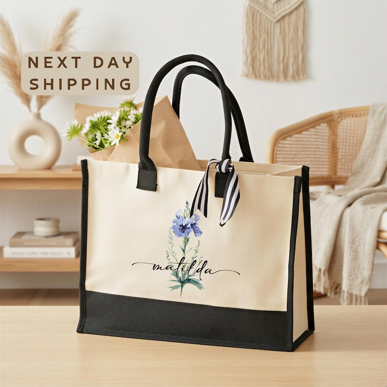 A beige tote bag with a floral design and the word "mala" printed on it, placed on a wooden surface. In the background, there is a beige wall with a hanging plant and a wooden chair. A sign that says "next day shipping" is visible in the top left corner of the image.