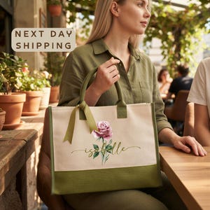 A woman is sitting at a table in an outdoor setting, holding a large tote bag with a floral design and the word "Isabella" printed on it.
