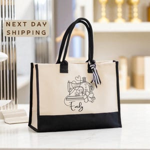 May include: Cream-colored tote bag with black accents and handles. The bag features a black graphic of a sewing machine, flowers, and the name "Emily." The words "Next Day Shipping" are in a brown rectangle in the upper left corner.