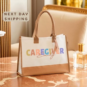 May include: A white tote bag with brown accents and handles. The bag features the word "CAREGIVER" in colorful letters, with "Emily" in cursive below. The bag is on a marble surface.
