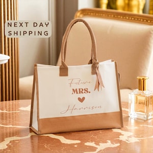 May include: A white tote bag with brown accents and handles. The bag features the text "Future MRS. Harrison" with a heart symbol. A ribbon is tied to the handle. The bag is sitting on a marble surface.