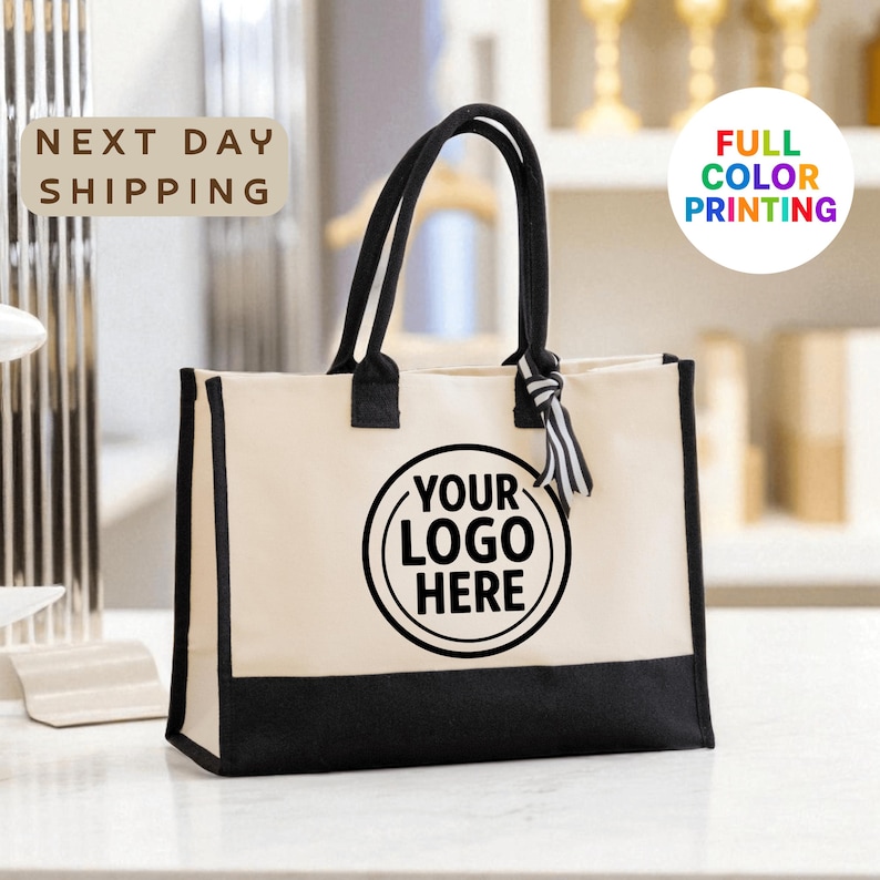 A beige tote bag with a black handle and a black and white striped ribbon tied around it, placed on a white surface.