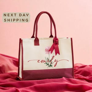 A white tote bag with a red ribbon and a pink flower design, sitting on a red fabric background with the text "Next day shipping" displayed above it.