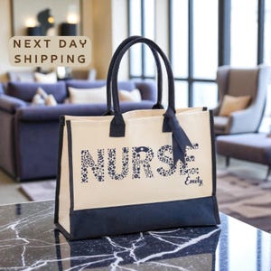 May include: Cream-colored tote bag with navy blue accents. The word "NURSE" is printed in navy blue, with floral and medical-themed designs within the letters. The name "Emily" is printed below the word "NURSE". The bag has navy blue handles.