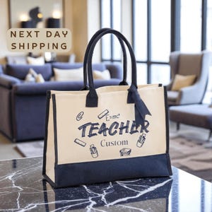 May include: Cream-colored tote bag with black handles and trim. The bag features the word "TEACHER" in a handwritten font, along with the word "Custom" and various school-related doodles. A dark blue ribbon is tied to one of the handles.