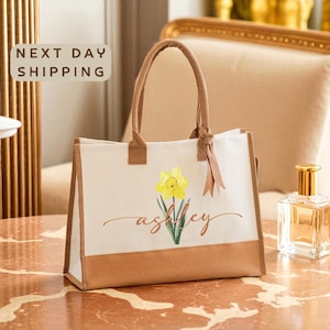A white tote bag with a yellow flower design and the name "Ashley" printed on it, placed on a marble table.