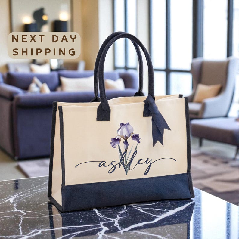A beige tote bag with a floral design and the name "Ashley" printed on it, placed on a marble countertop.