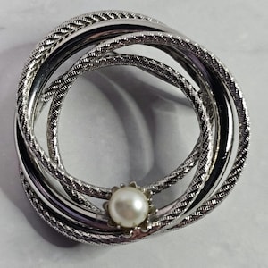 May include: Silver-toned brooch with three intertwined rings, each with a textured rope-like design. A central pearl-like bead is set in a scalloped, silver-toned setting. The brooch has a classic, elegant style.