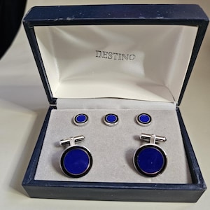 May include: A set of cufflinks and shirt studs in a navy blue box. The cufflinks and studs have a silver-tone metal finish with a dark blue center and a black border. The box is navy blue with the word "DESTINO" printed on the inside.