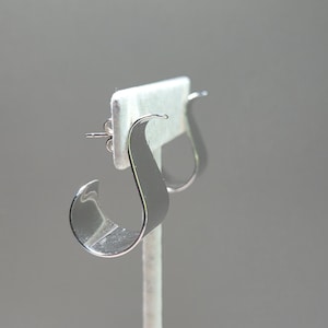 May include: A pair of silver-toned hoop earrings with a smooth, reflective surface and a distinct S-shaped curve. The earrings feature a post-back closure mechanism, providing a secure fit. The design is characterized by its open, fluid silhouette.