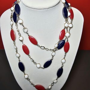 May include: A multi-strand necklace featuring alternating oval-shaped beads in navy blue, bright red, and white. The beads are connected by thin silver-toned metal links, creating a layered design with a repeating pattern of colored and white accents.